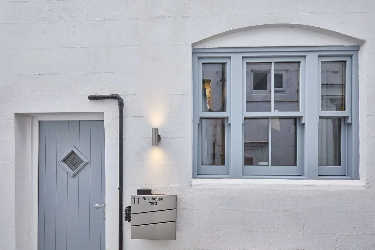 Una fachada clara con una puerta gris, una ventana y una luz de pared.