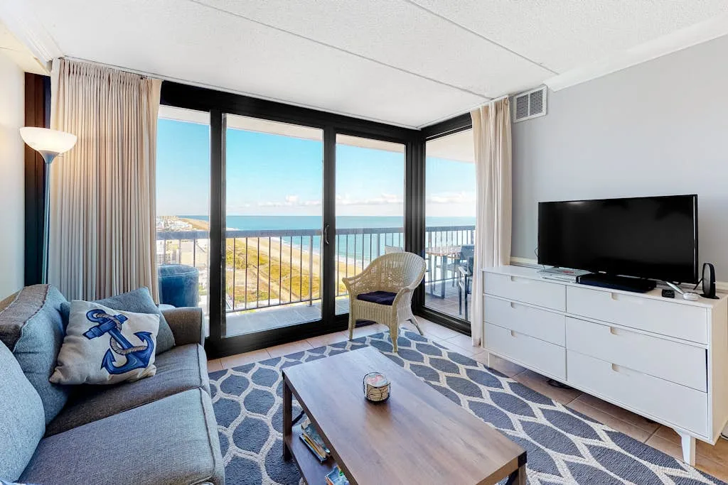 A bright living room with a seating area, television, and ocean views through large windows.