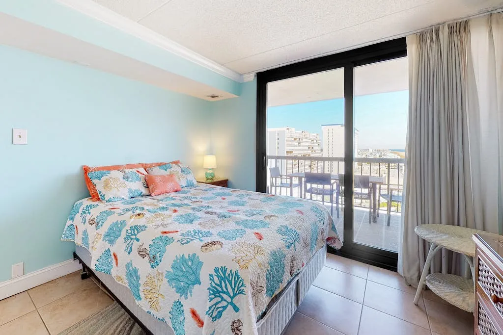 A bedroom with a patterned bedspread beside glass doors opening onto a balcony.