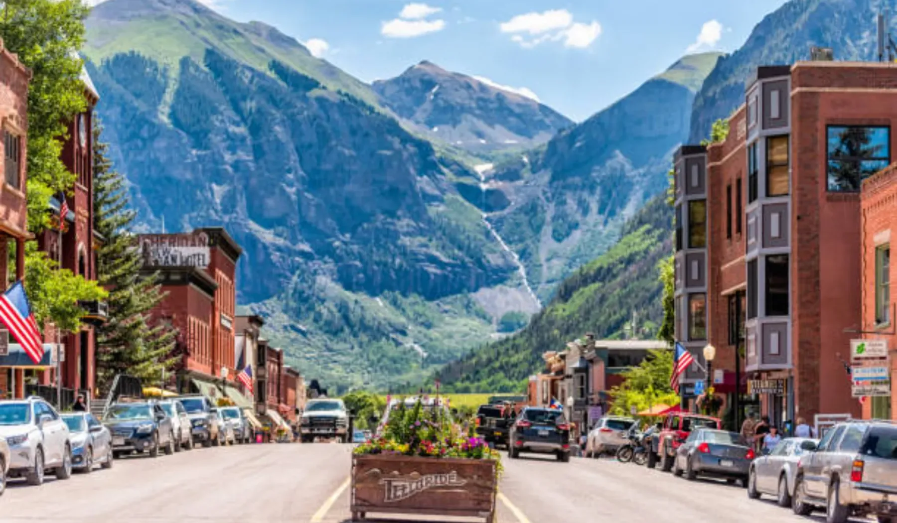 Una calle de un pueblo de montaña con vehículos estacionados y picos escarpados al fondo.
