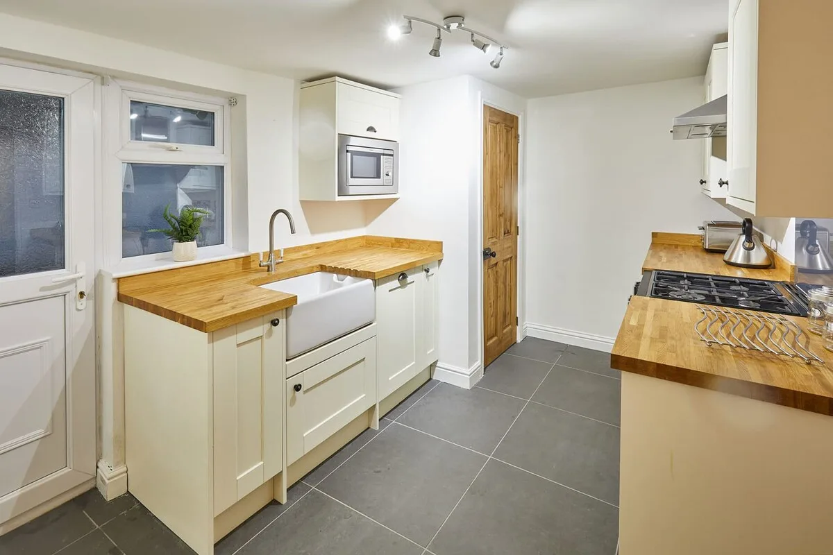 A bright kitchen with wooden countertops, white cabinets, and gray floor tiles.