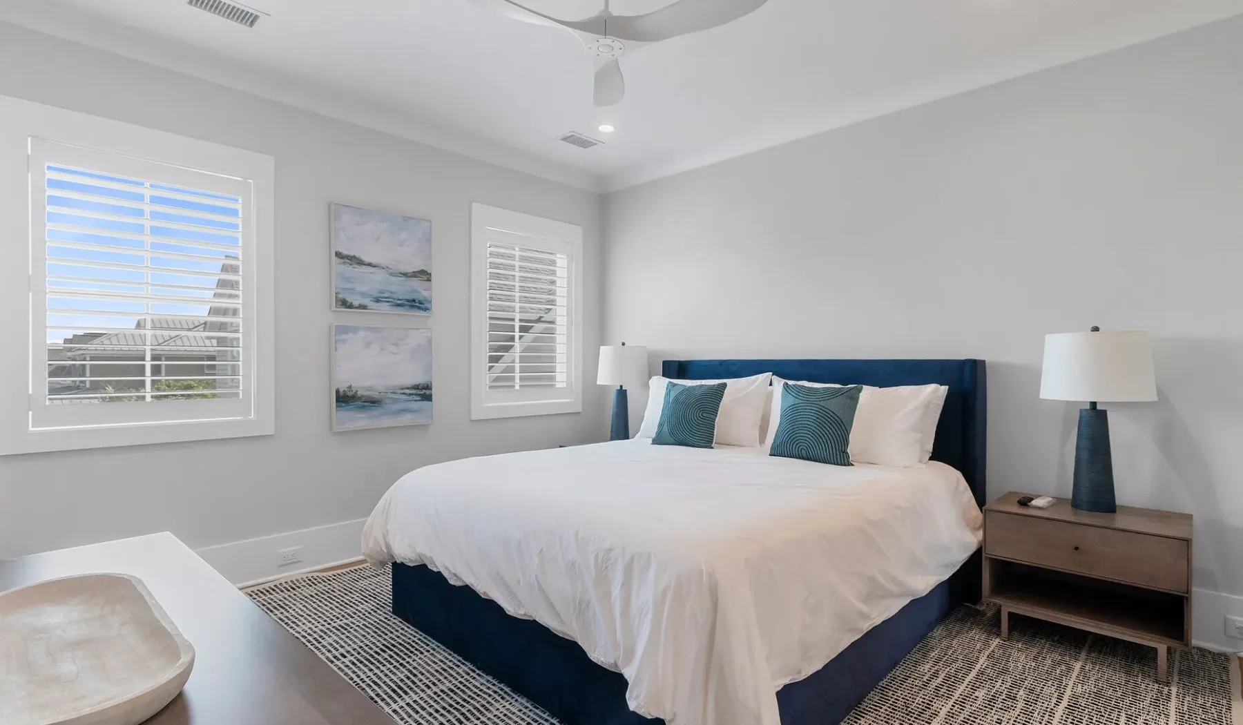 Un dormitorio luminoso con una cama, dos ventanas y cuadros enmarcados.