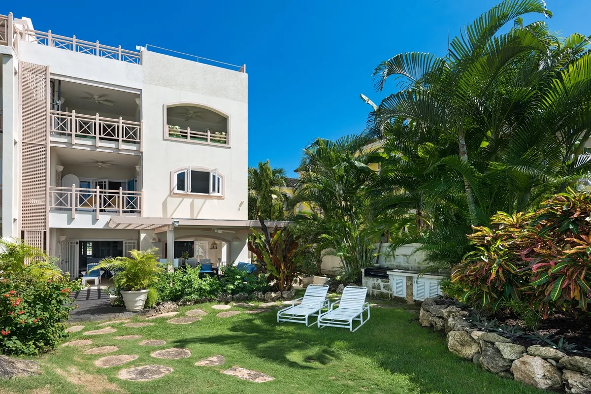 A garden patio with outdoor seating sits beside a white multi-story building and palm trees.