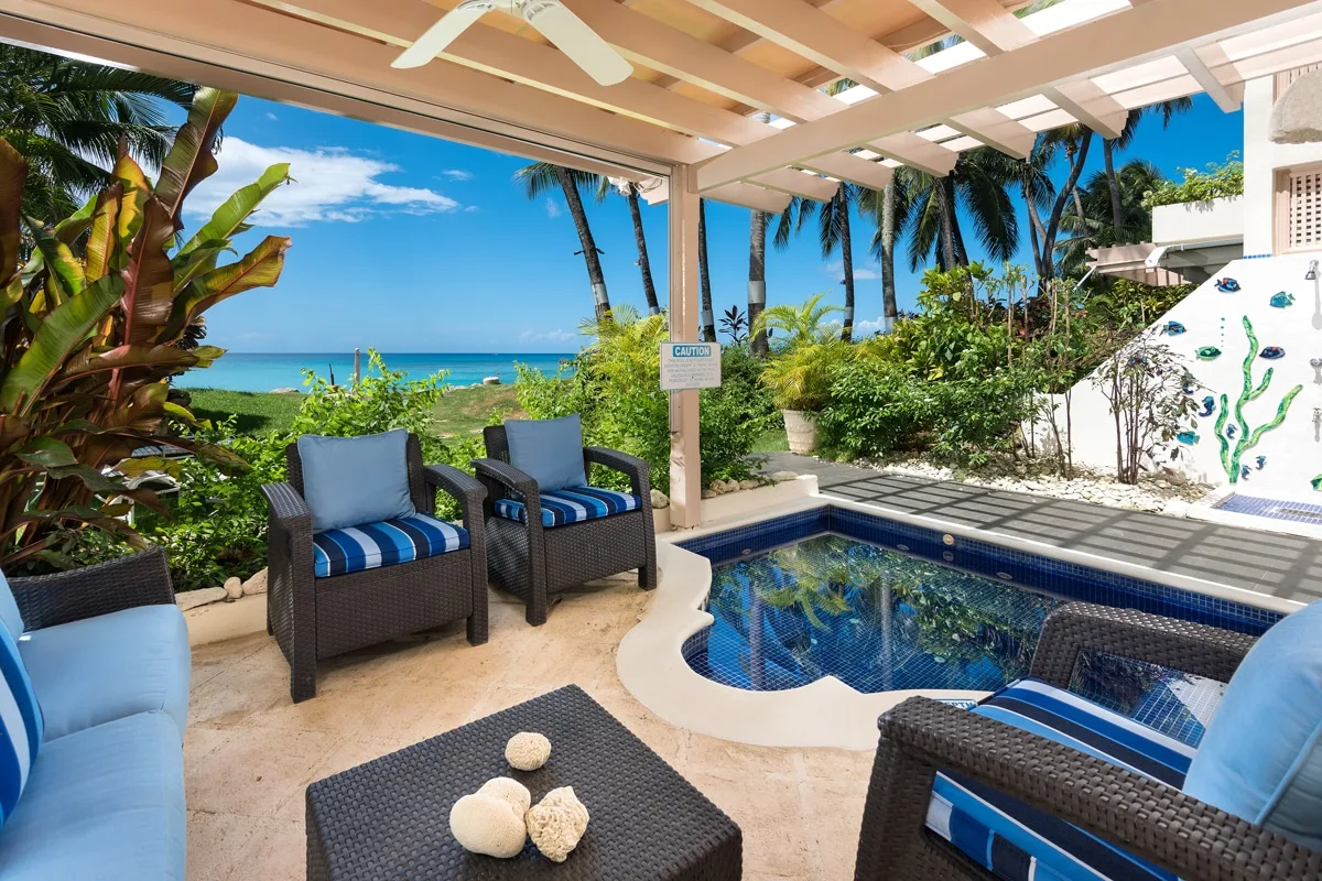 A poolside patio with cushioned seating overlooks tropical plants and the ocean.