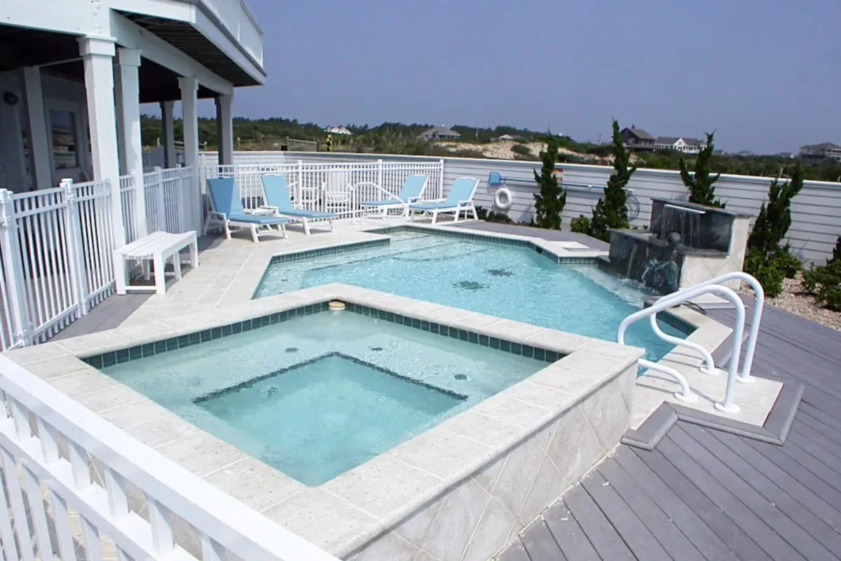An outdoor pool and raised spa sit on a deck beside a white-railed porch.