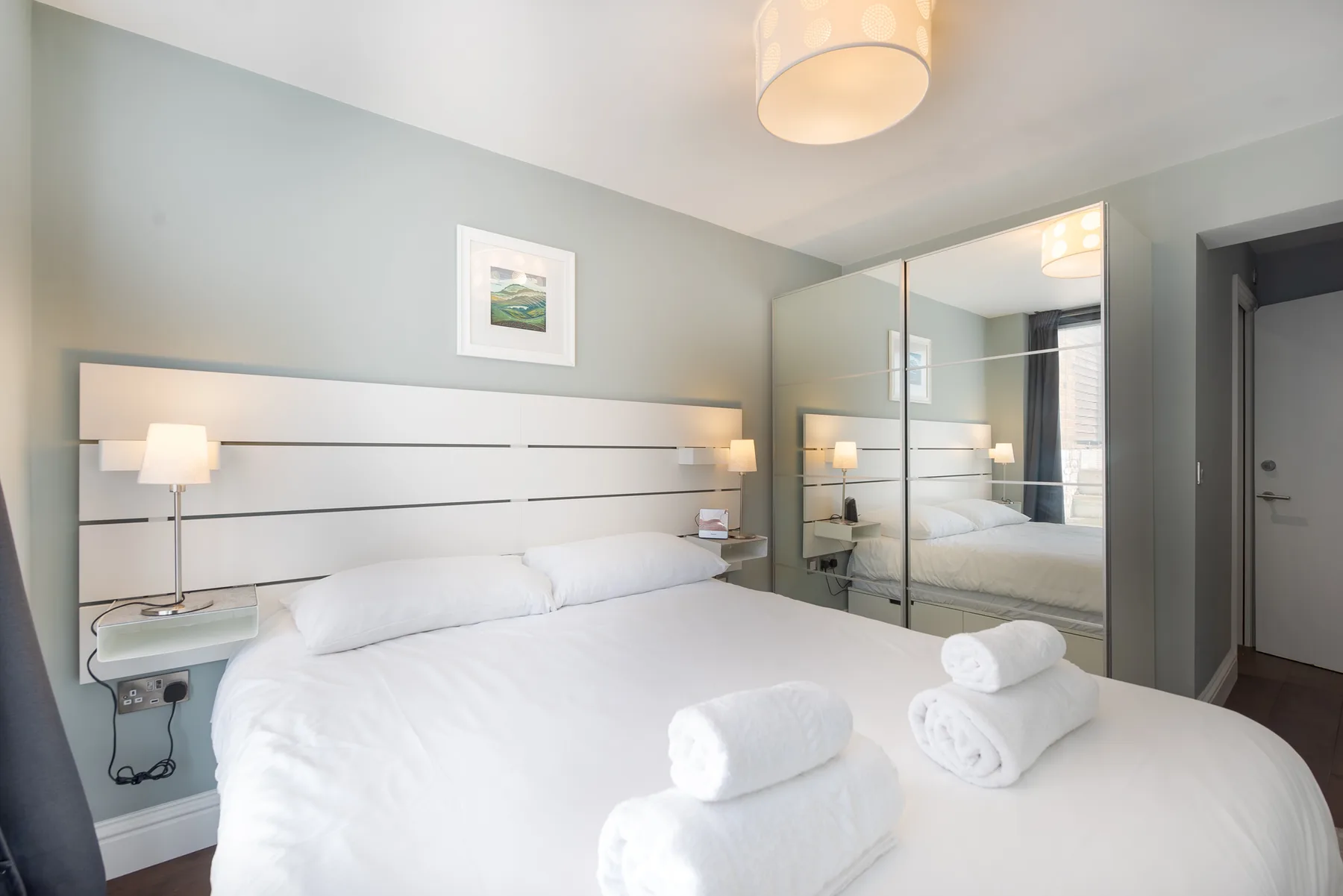 A bright bedroom with a large bed and mirrored wardrobe doors.