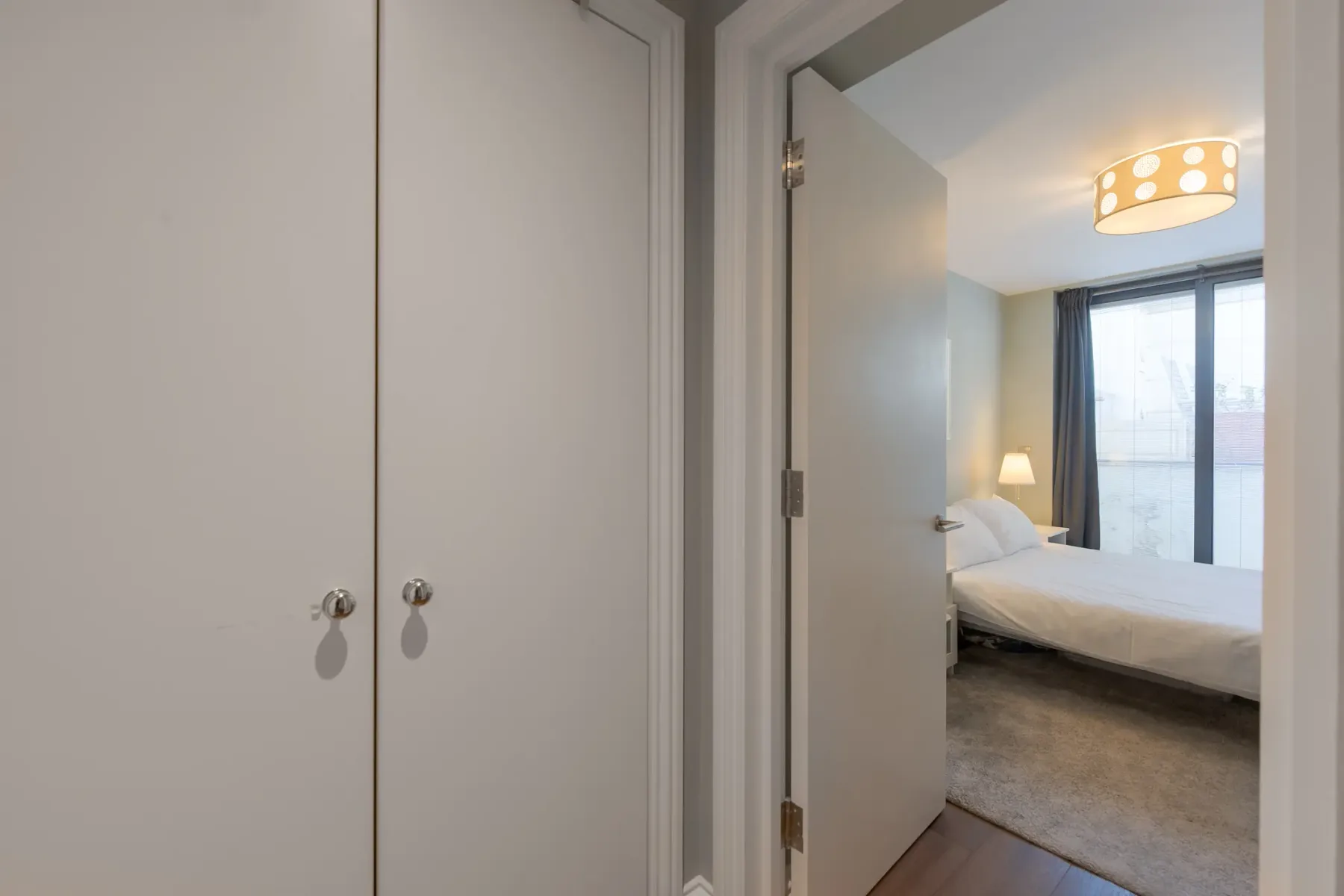 A bedroom with a bed and large windows, viewed through an open doorway.