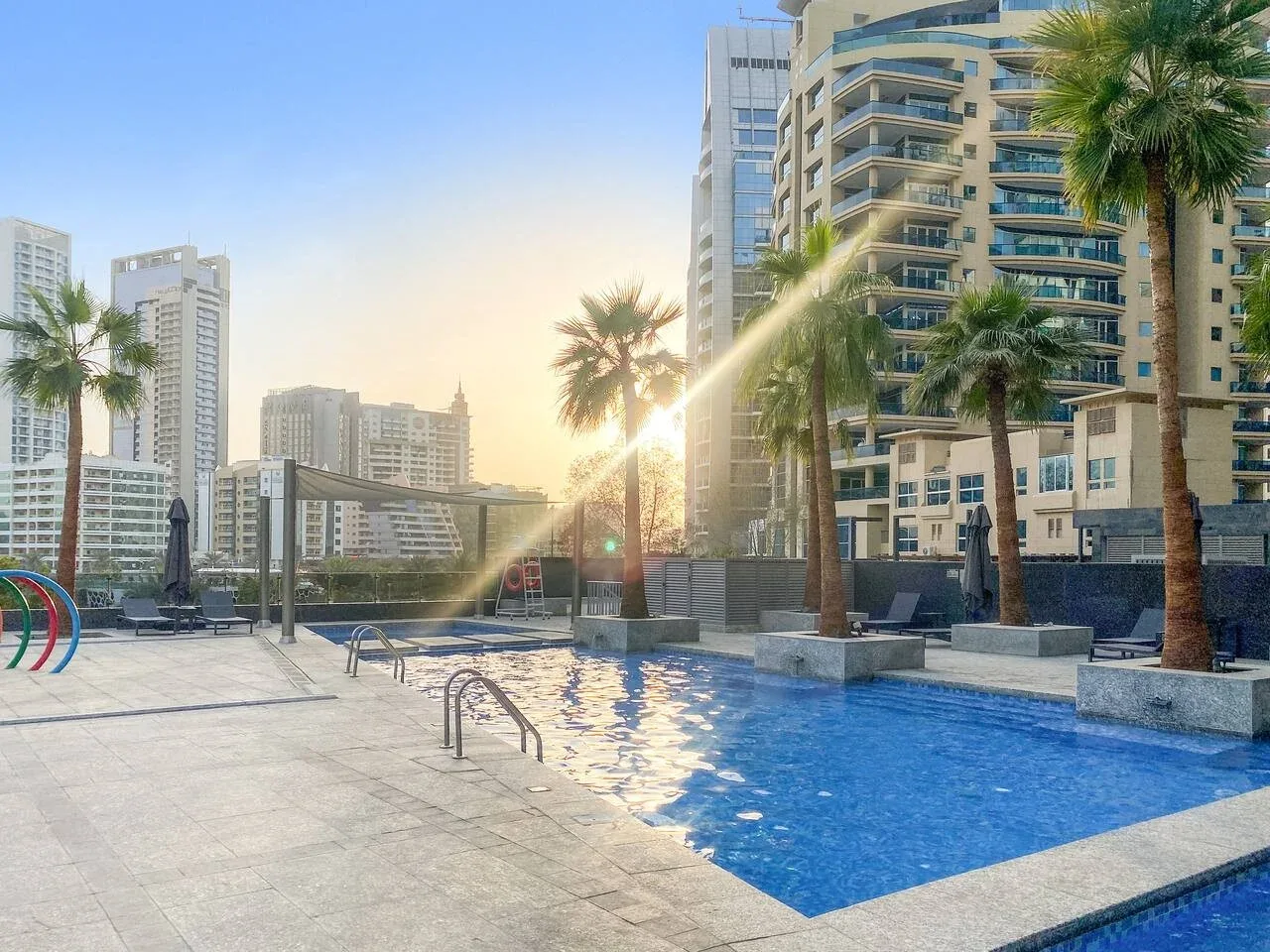 An outdoor swimming pool sits beside palm trees and high-rise buildings.