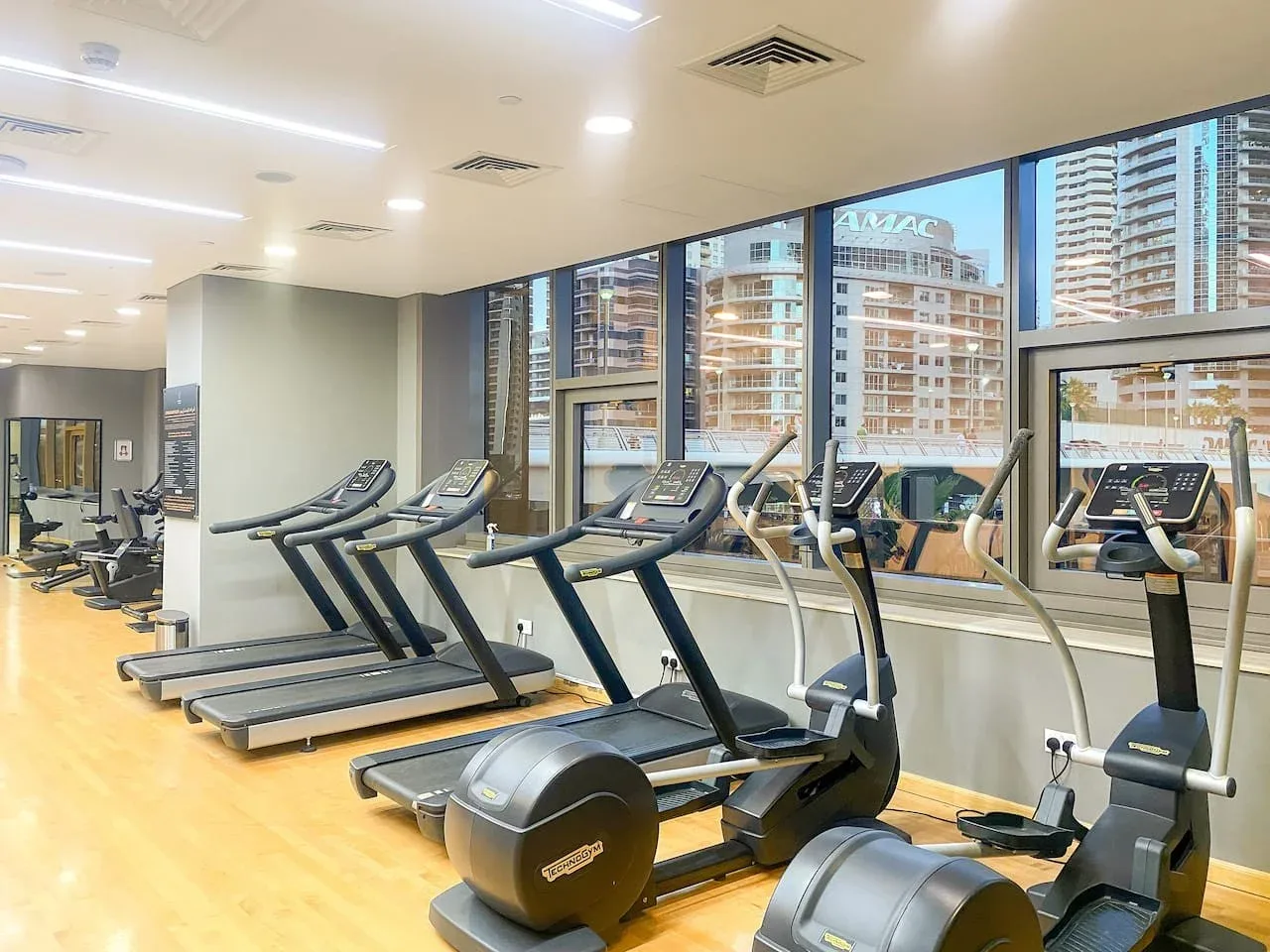 A gym with treadmills and exercise bikes beside large windows.