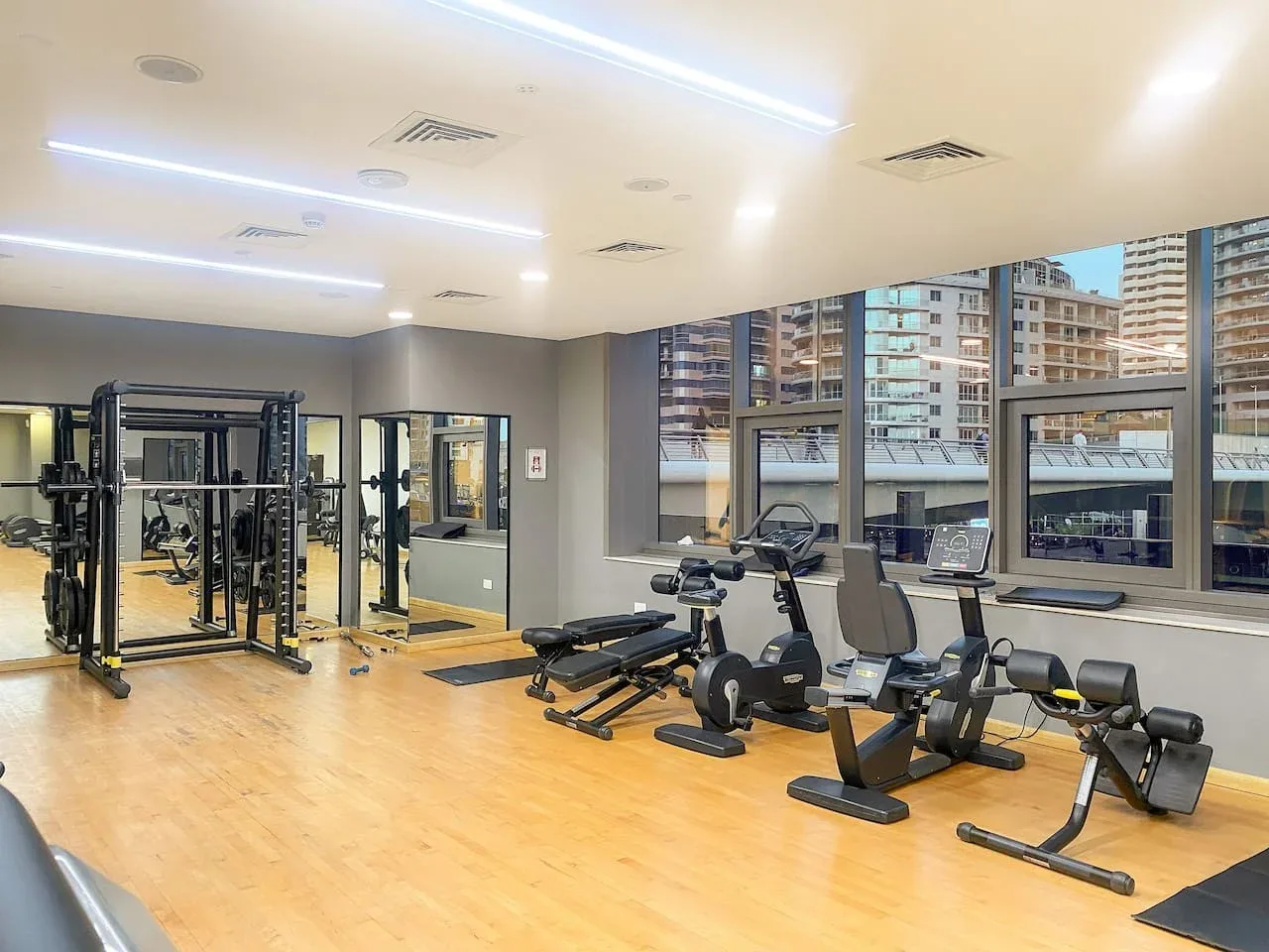 A fitness room with exercise machines and large windows overlooking nearby buildings.