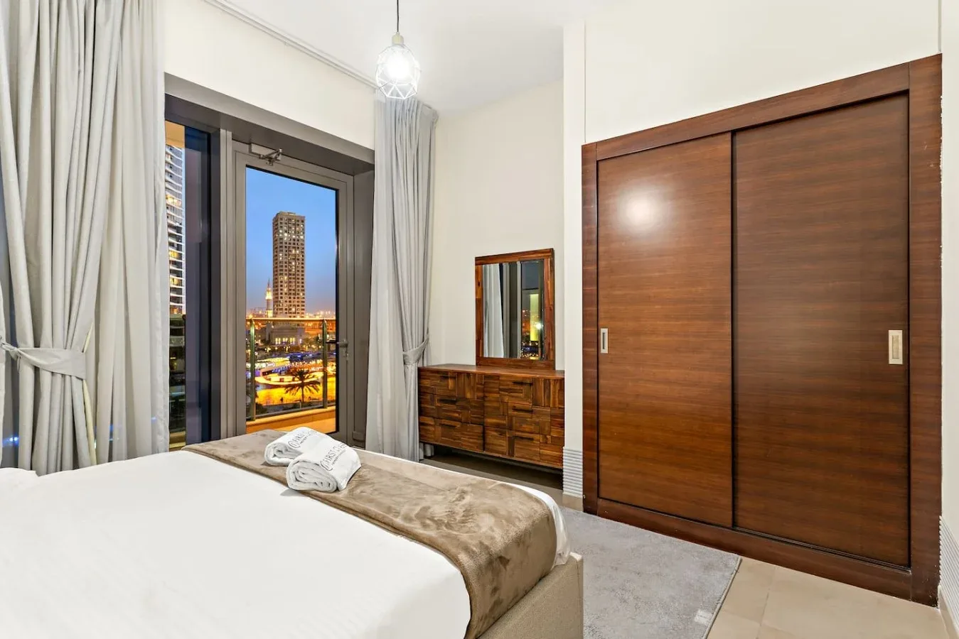 A bedroom with a large wooden wardrobe and a window overlooking illuminated buildings.