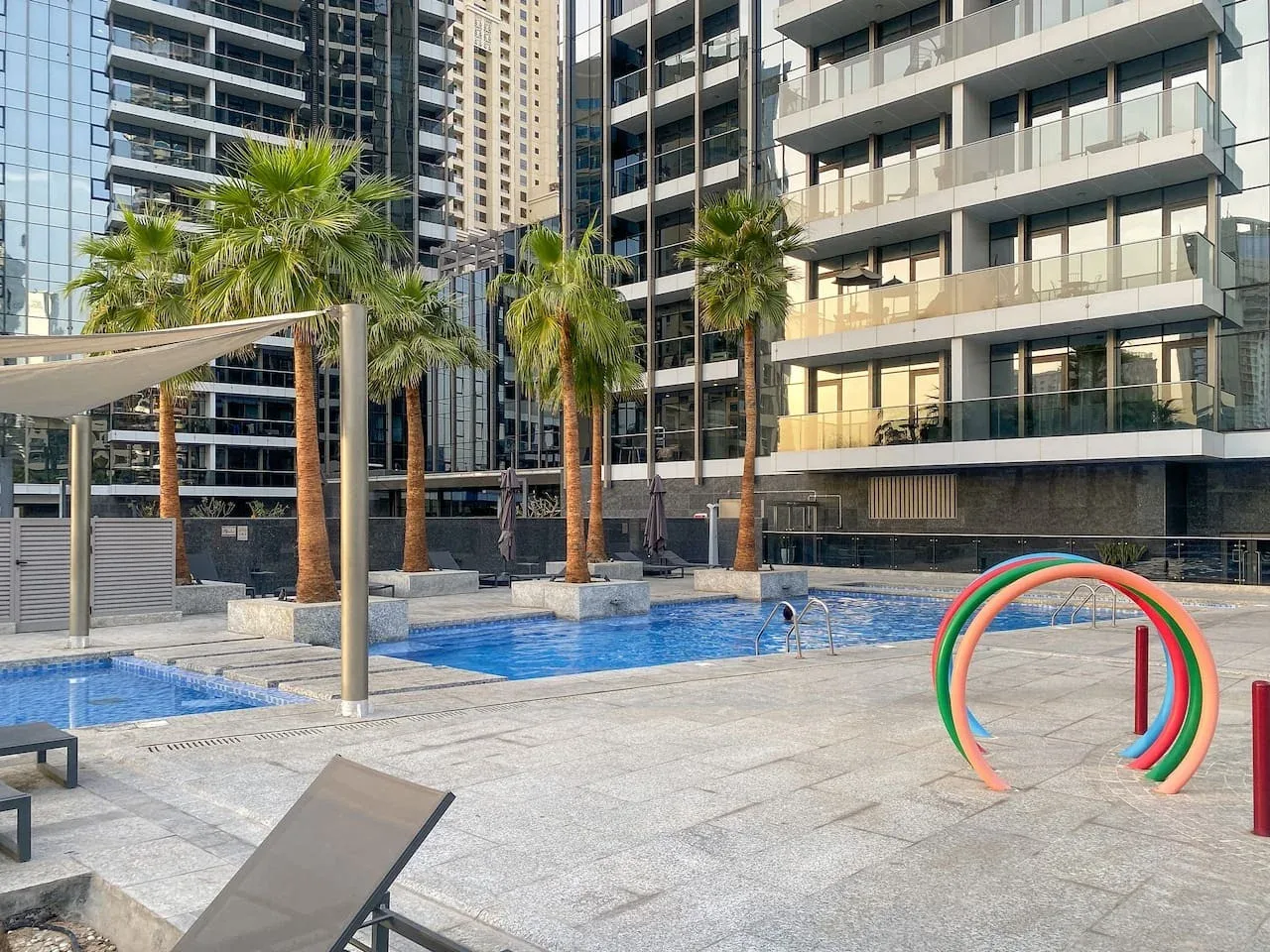 An outdoor swimming pool with palm trees and surrounding high-rise buildings.
