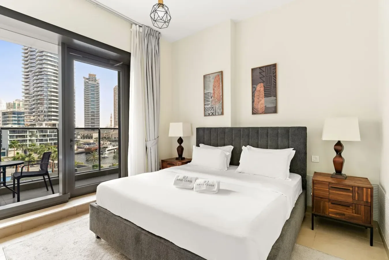 A bright bedroom with a neatly made bed beside large windows overlooking the city.