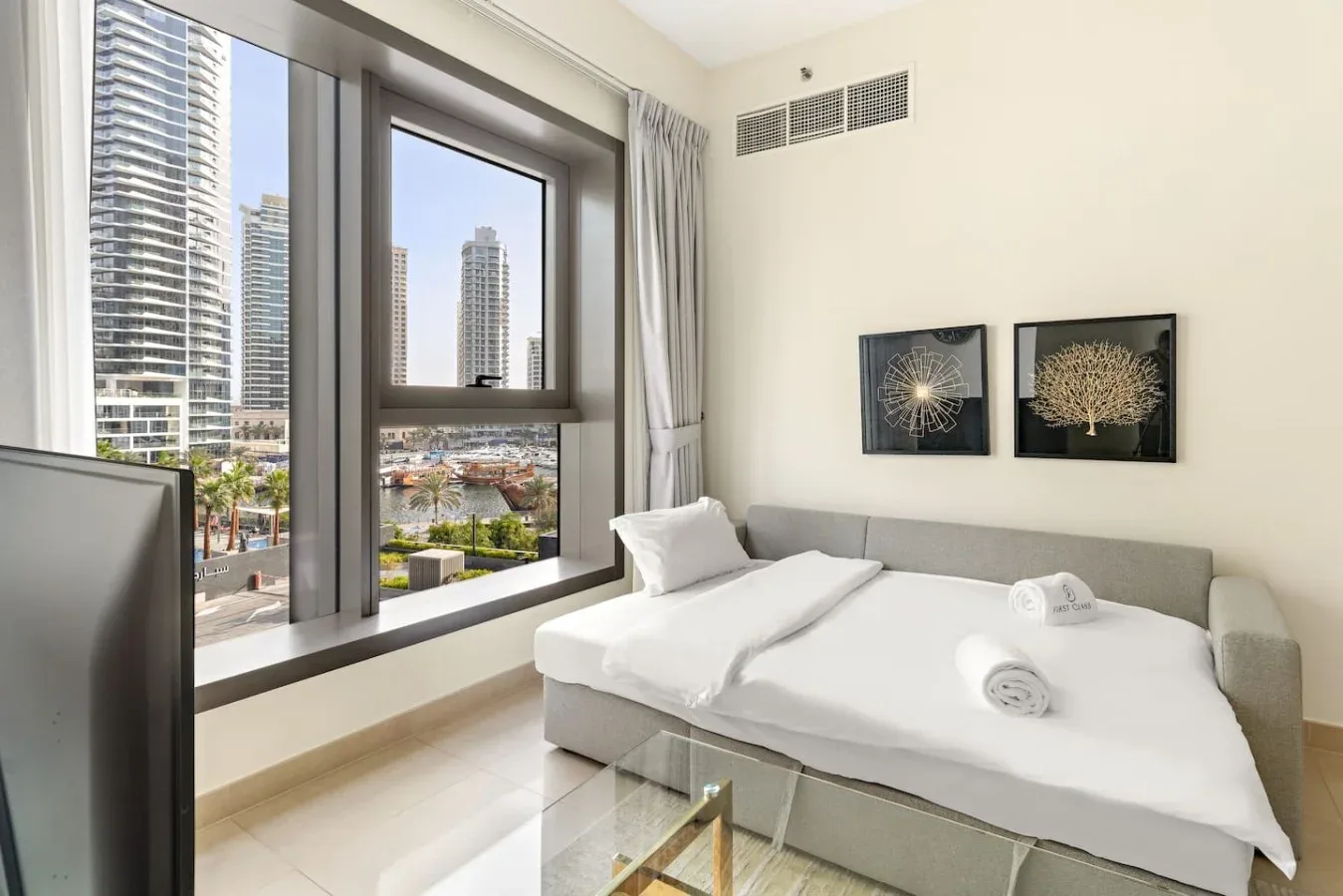 A bright bedroom with a bed beside large windows overlooking nearby buildings.