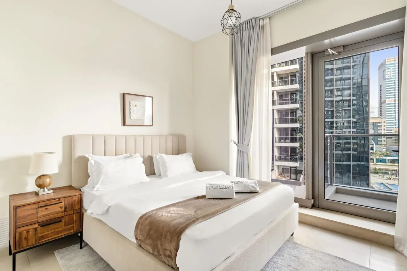 A bright bedroom with a neatly made bed beside large windows overlooking nearby buildings.