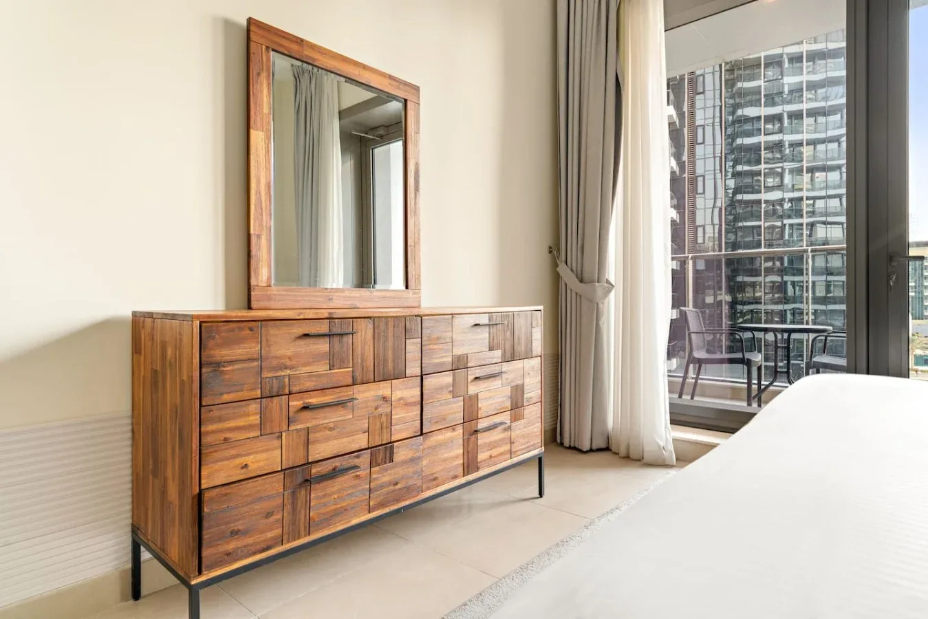 A bedroom with a wooden dresser and mirror beside a large window.