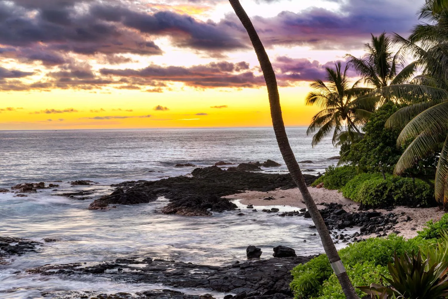 Una costa tropical rocosa se encuentra con el océano bajo un cielo colorido al atardecer.