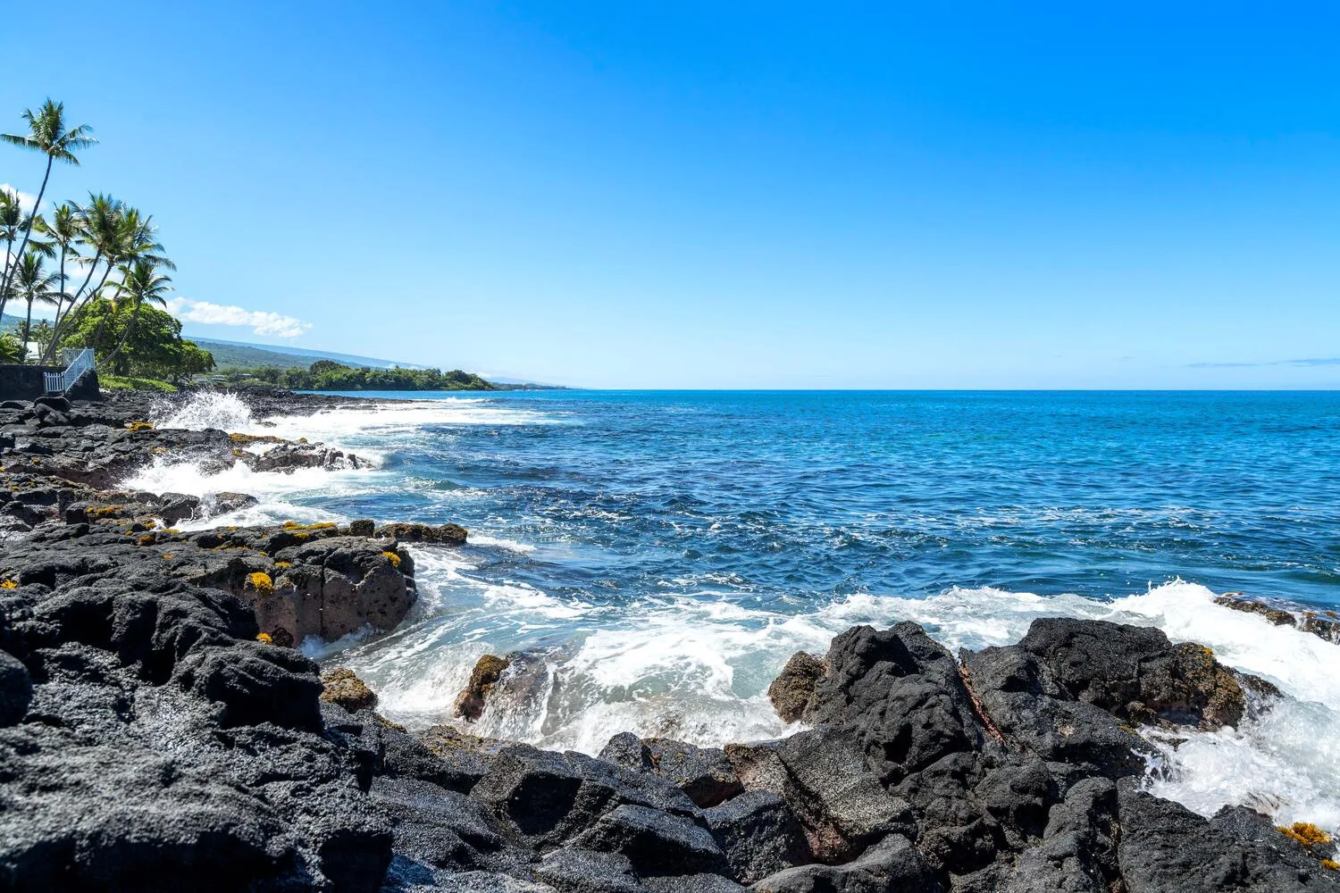 Una costa rocosa con olas rompiendo contra rocas oscuras bajo un cielo azul despejado.