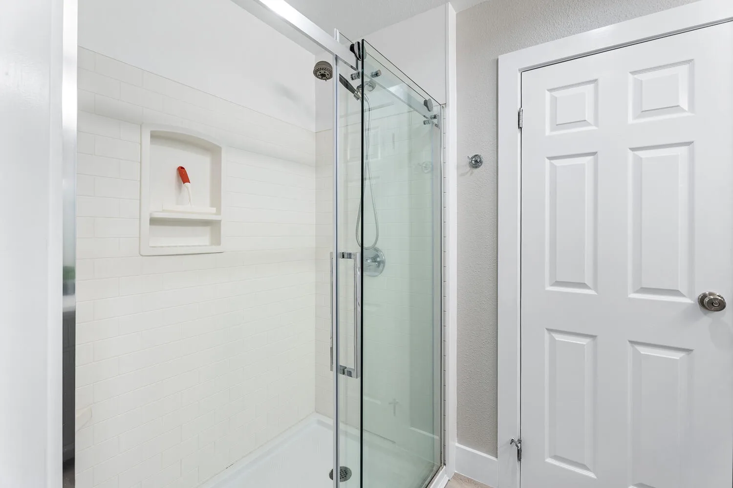 Un baño luminoso con una ducha de vidrio junto a una puerta blanca con paneles.