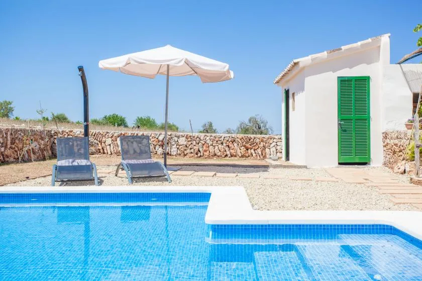 An outdoor swimming pool beside a white building, with seating under a sun umbrella.