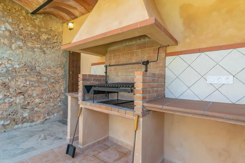 A built-in brick barbecue stands beneath a hood in a rustic indoor cooking area.