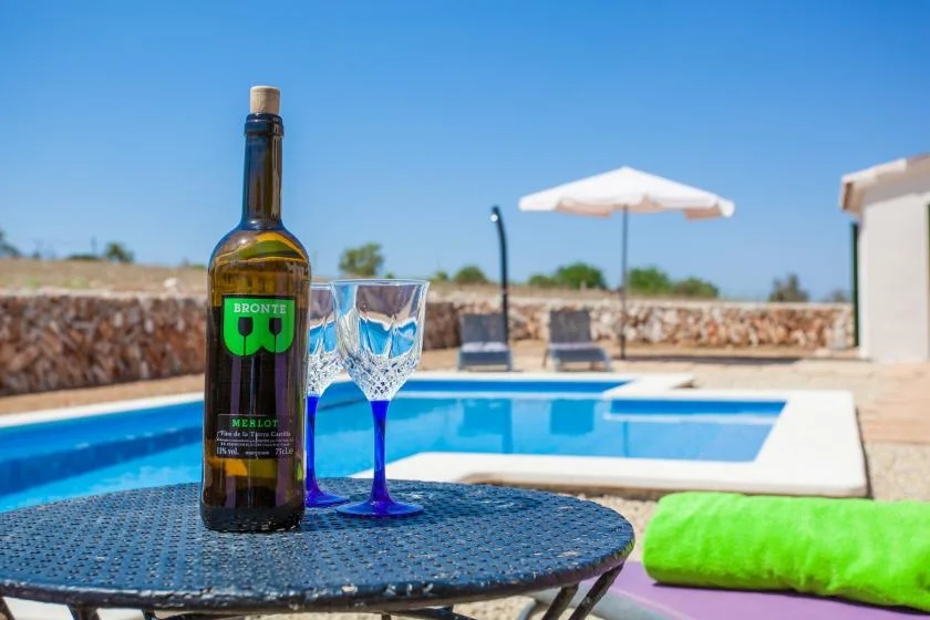 A bottle and glass sit beside a swimming pool with a sun umbrella in the background.