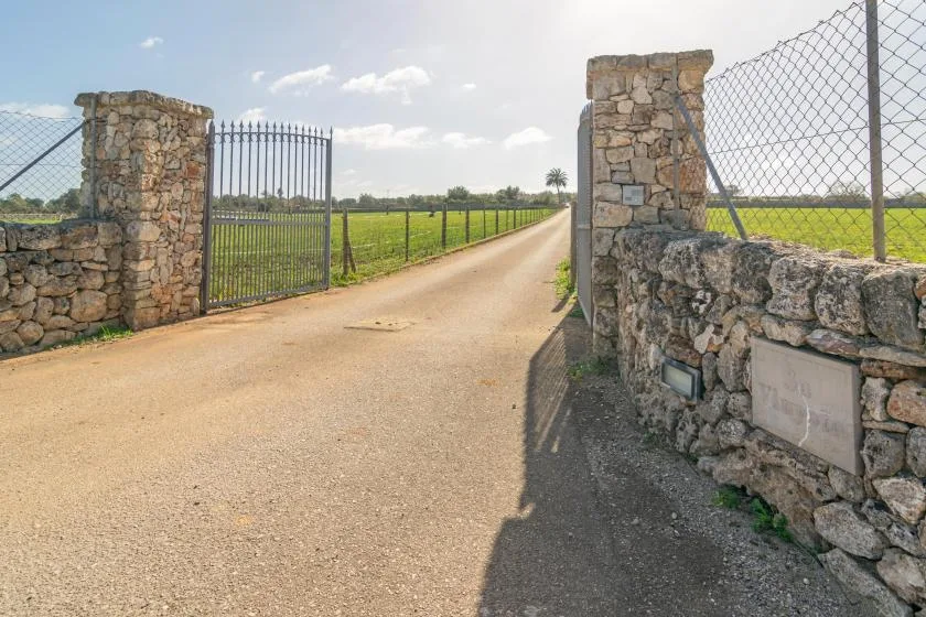 A rural entrance road passes through a metal gate between stone pillars and fencing.