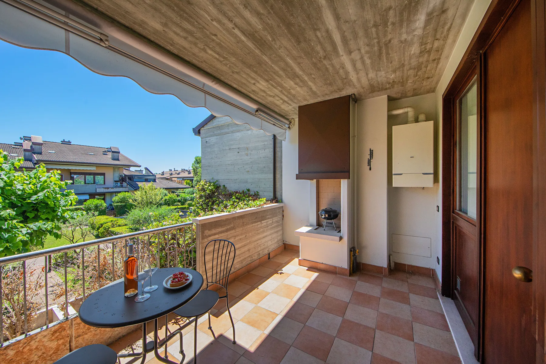 A covered balcony with a small table and chairs overlooks a green residential area.