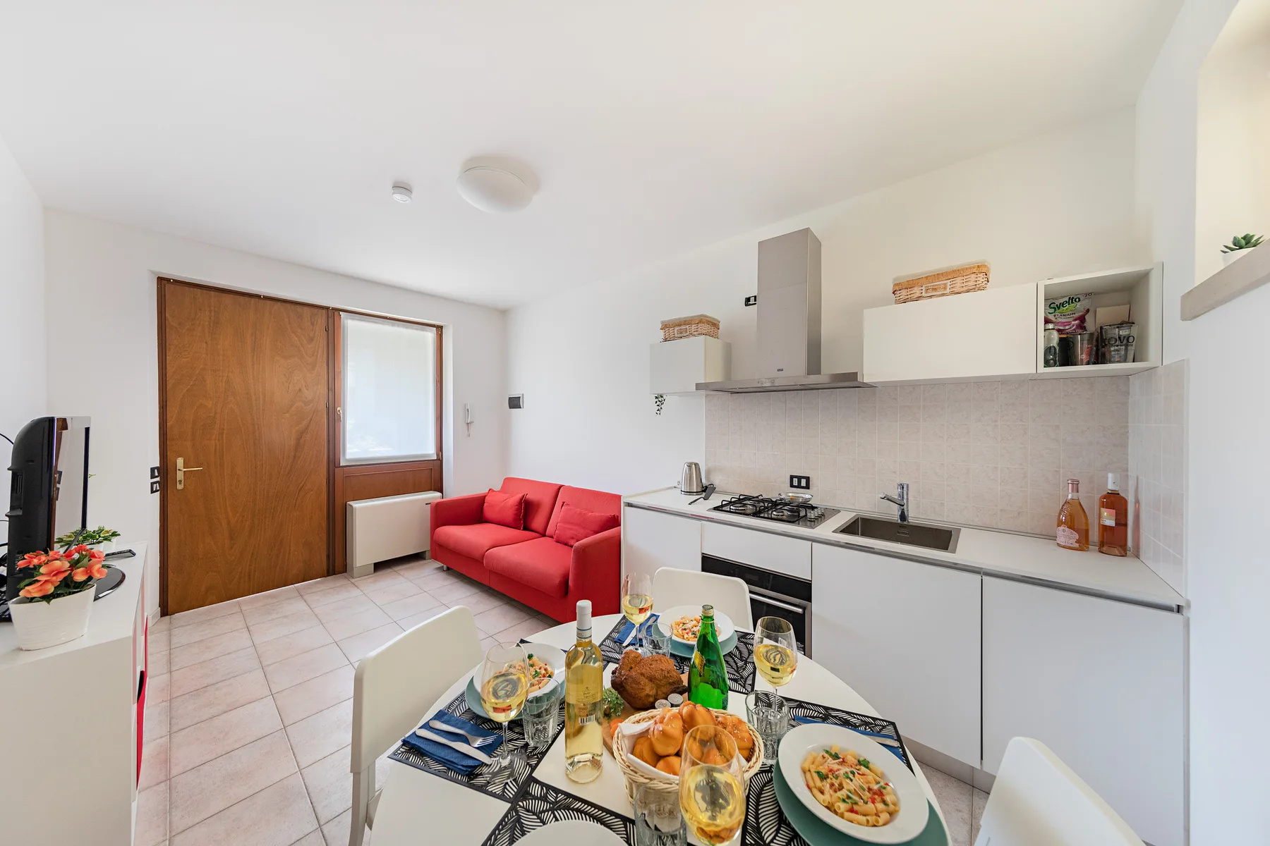 A bright open-plan room combines a red sofa, dining table, and compact kitchen.