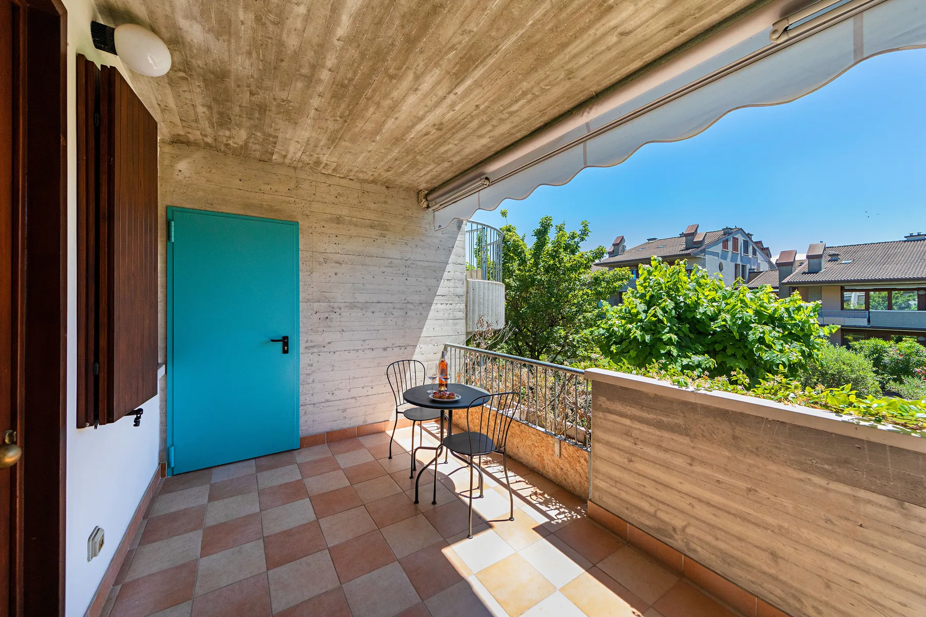 A covered balcony has a small table and seating overlooking leafy trees.