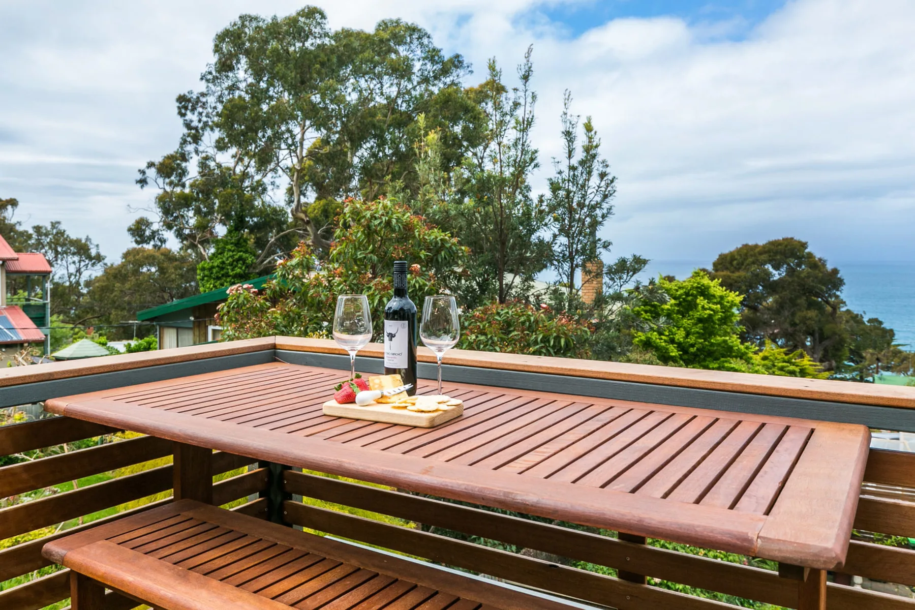 A wooden outdoor dining table with wine and glasses overlooks a leafy garden.