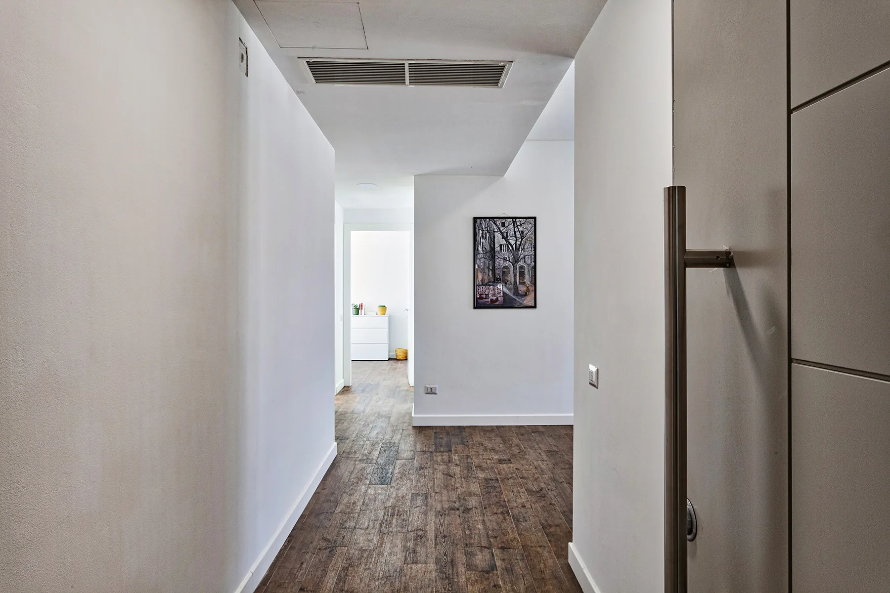 Un pasillo estrecho con suelo de aspecto madera y una obra enmarcada en la pared.