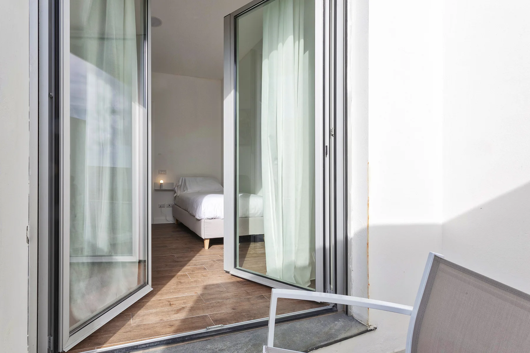 Una puerta corrediza de vidrio abierta da a un dormitorio con una cama y piso de madera.