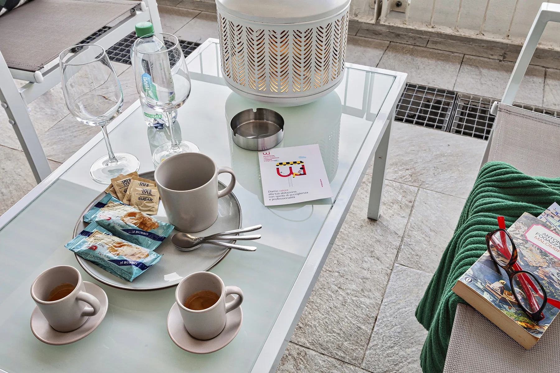Una mesa de centro de vidrio sostiene tazas, aperitivos y material de lectura junto a asientos al aire libre.