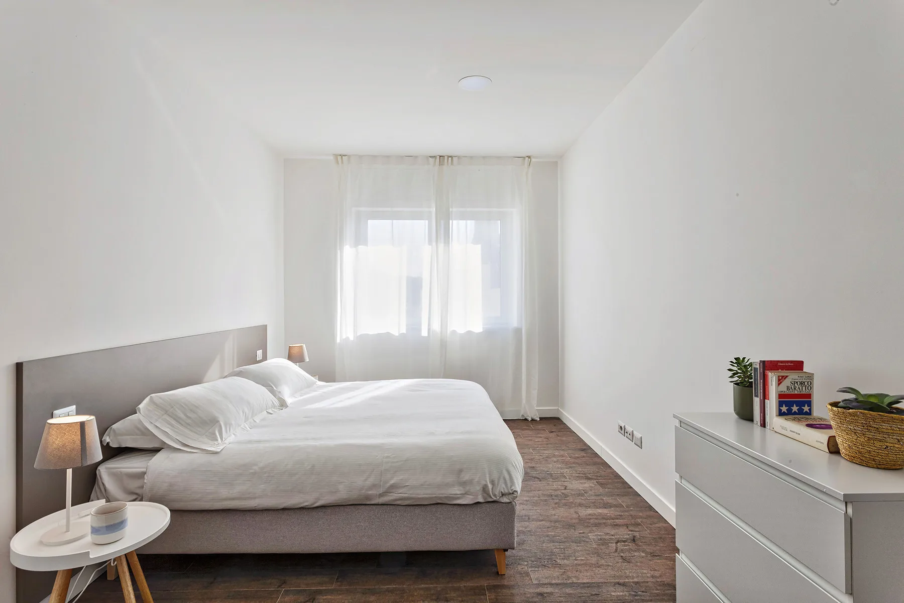 Un dormitorio luminoso con una cama, una lámpara de noche y una ventana con cortinas translúcidas.