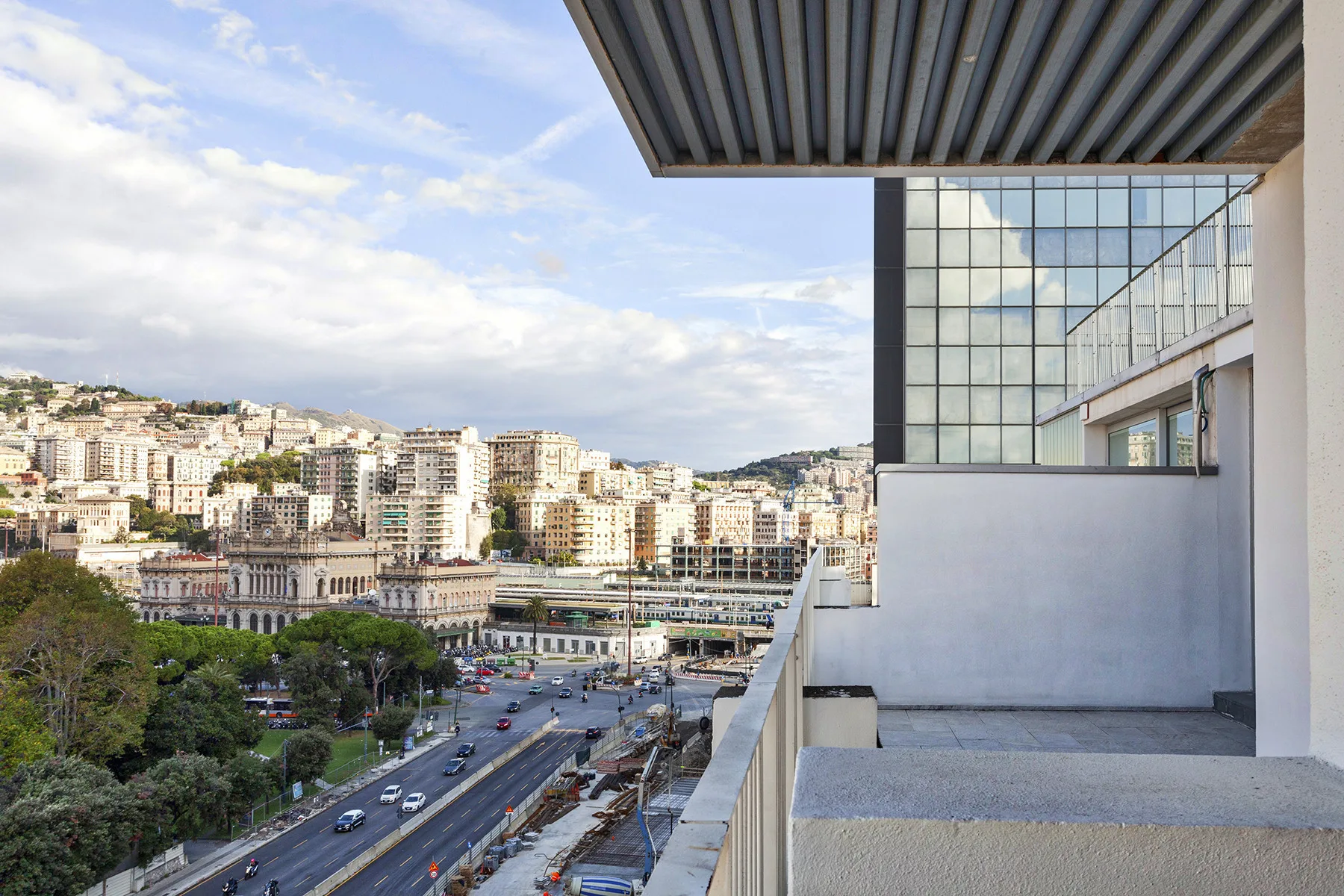 Un balcón da a una calle de la ciudad, zonas verdes y edificios en una colina.