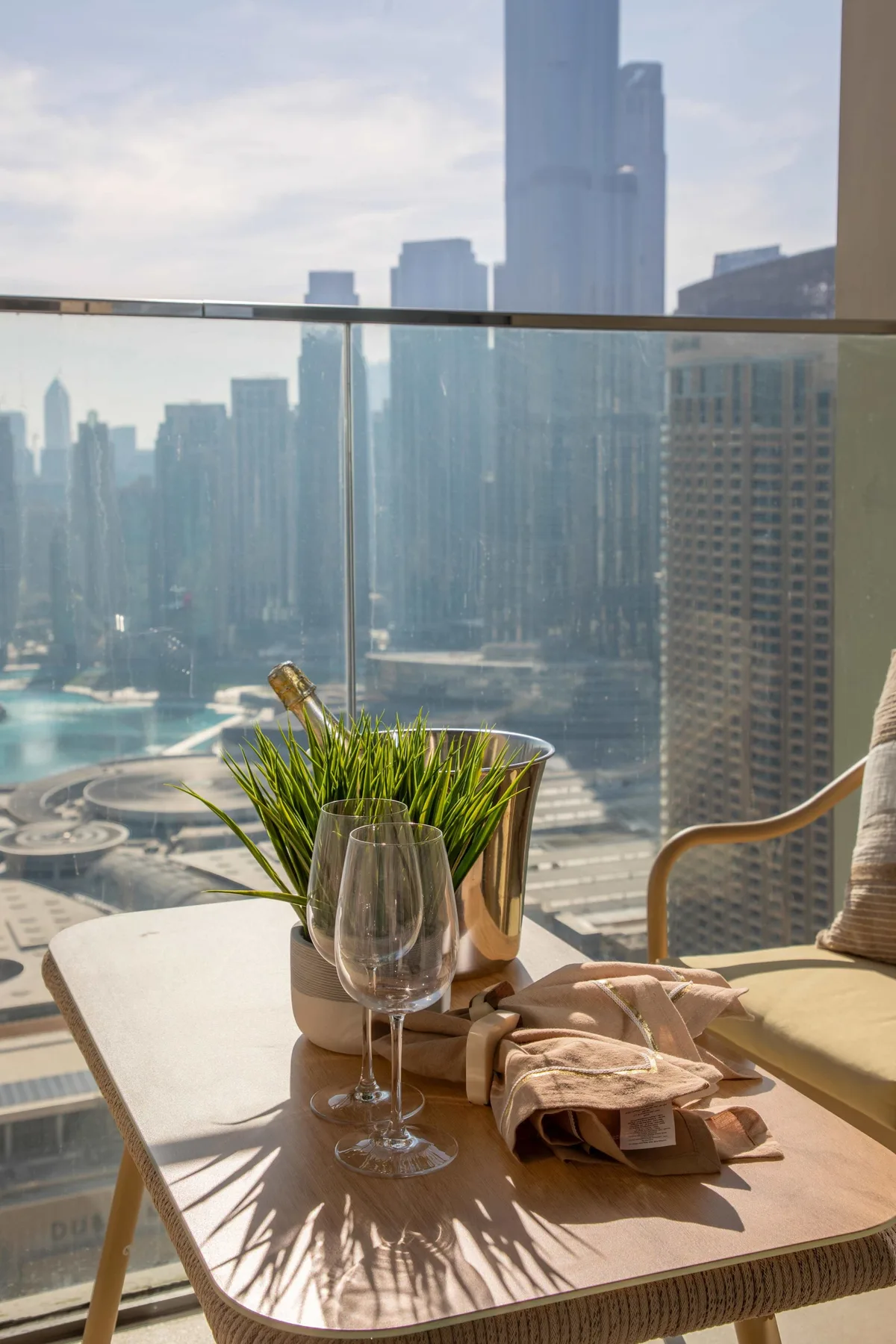 A balcony table with glasses and a potted plant overlooks a city skyline.