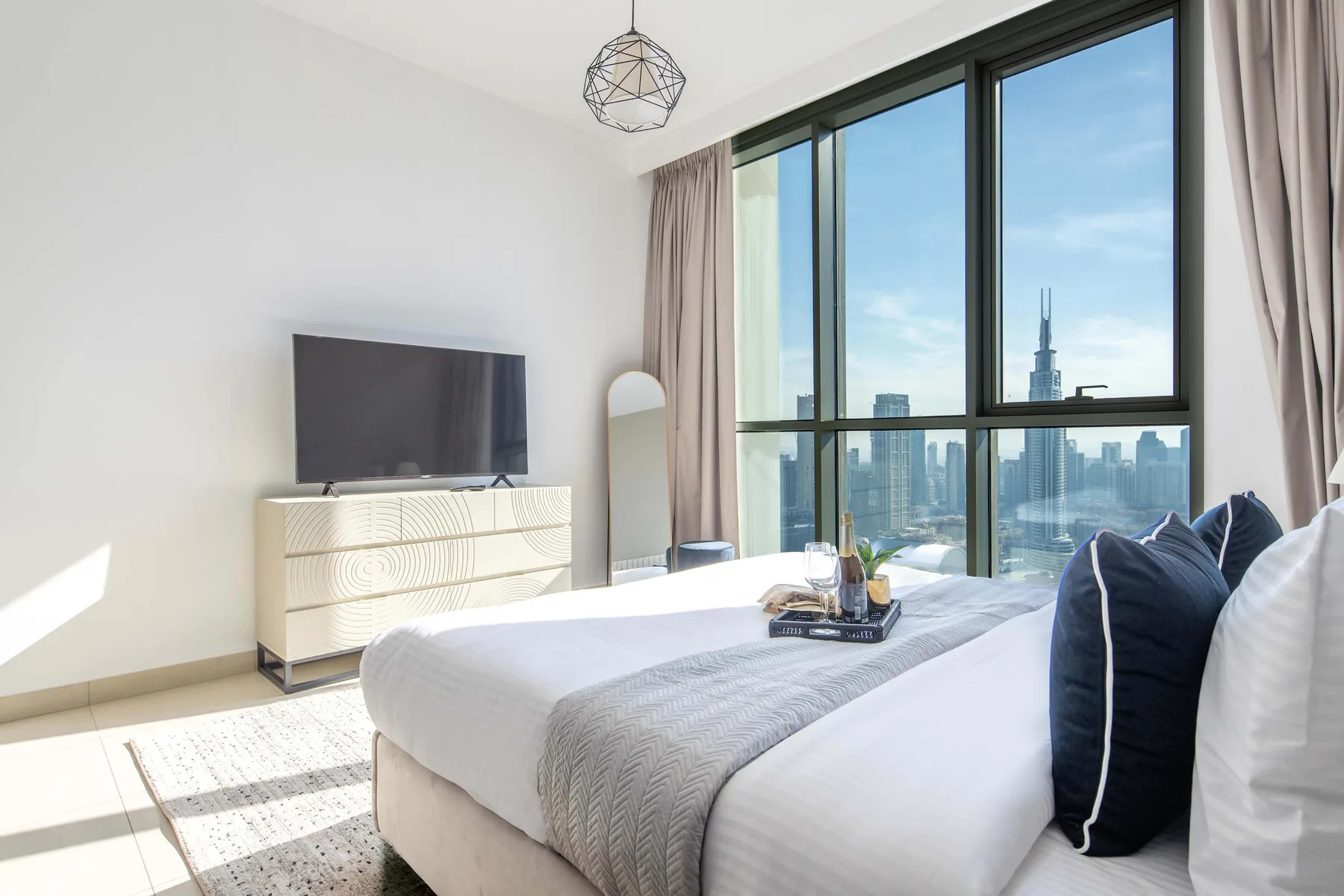 A bright bedroom with a neatly made bed beside large windows overlooking the city.