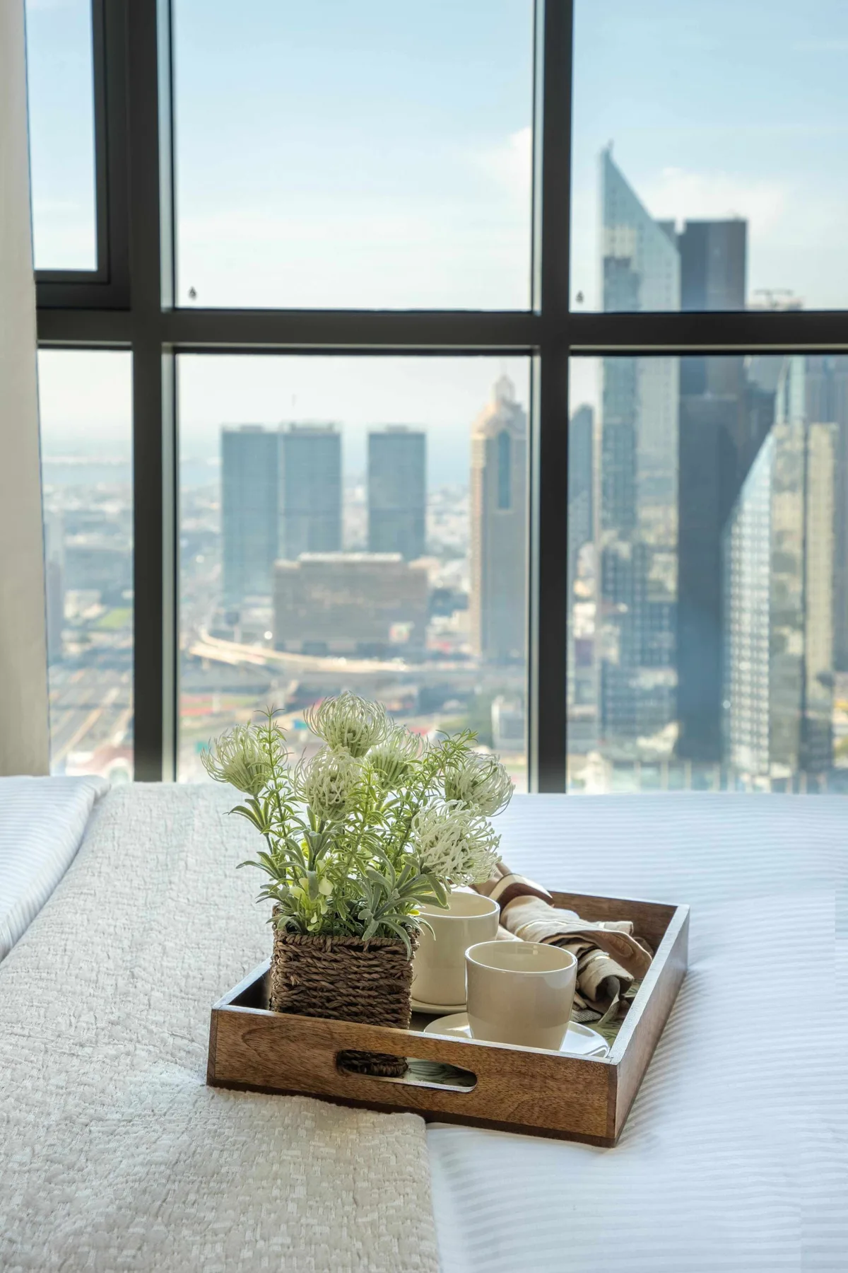 A bed with a wooden tray and flowers sits beside a large city-view window.
