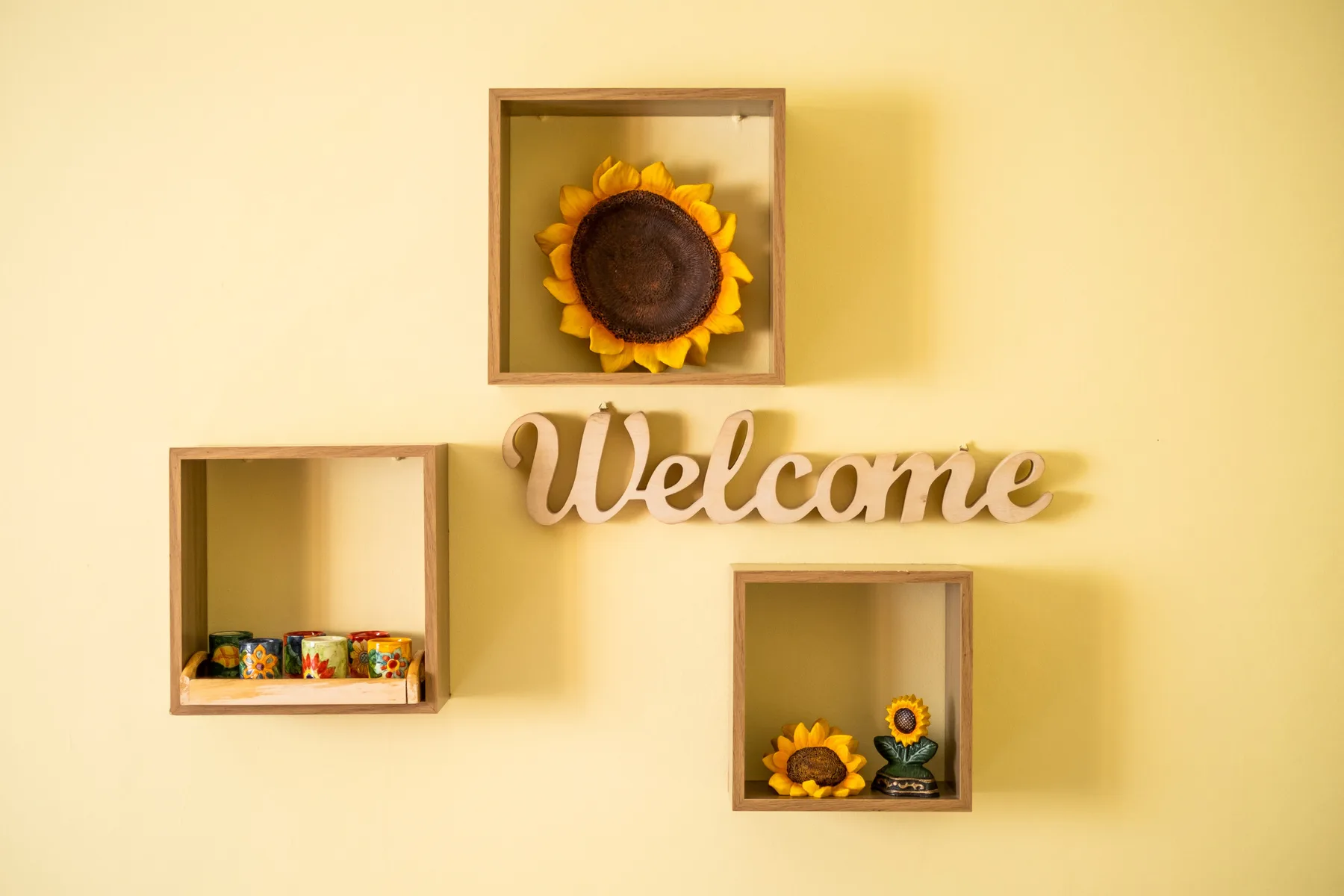 Una pared amarilla muestra un letrero decorativo de bienvenida y tres pequeños estantes.
