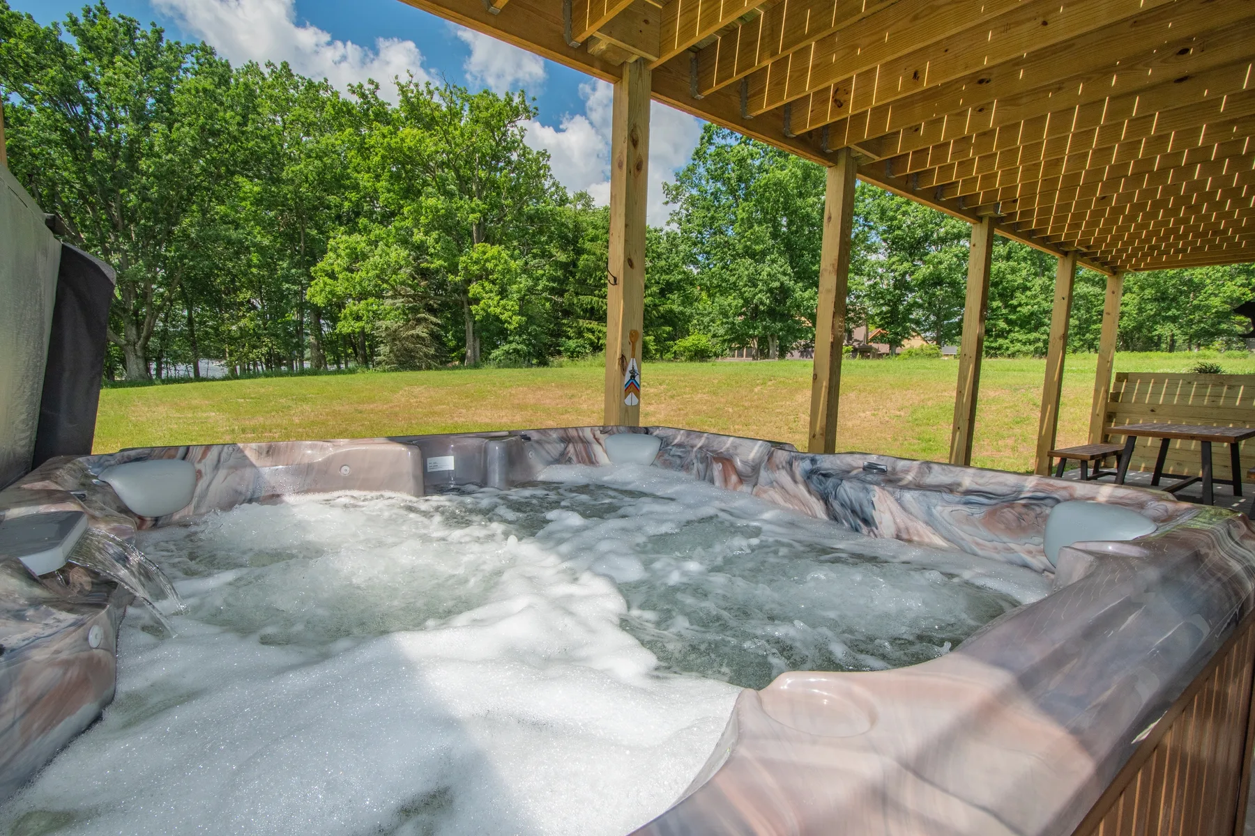 An outdoor hot tub sits beneath a wooden covered patio, overlooking a grassy yard.