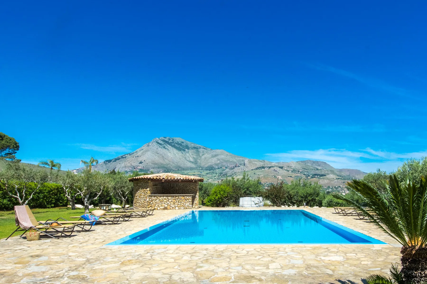 An outdoor swimming pool sits on a stone-paved terrace with mountains in the distance.