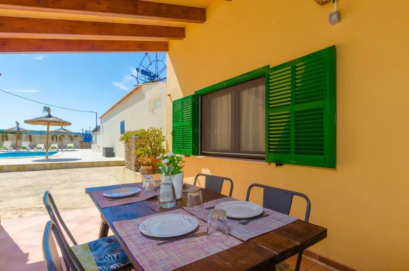 A dining table sits beneath a covered patio beside a window with green shutters.