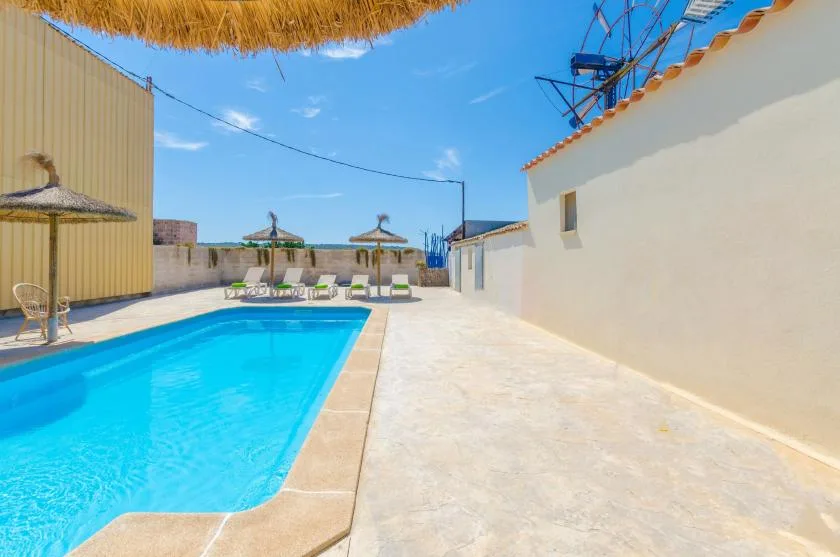 An outdoor swimming pool sits beside a sunny paved patio with lounge seating.
