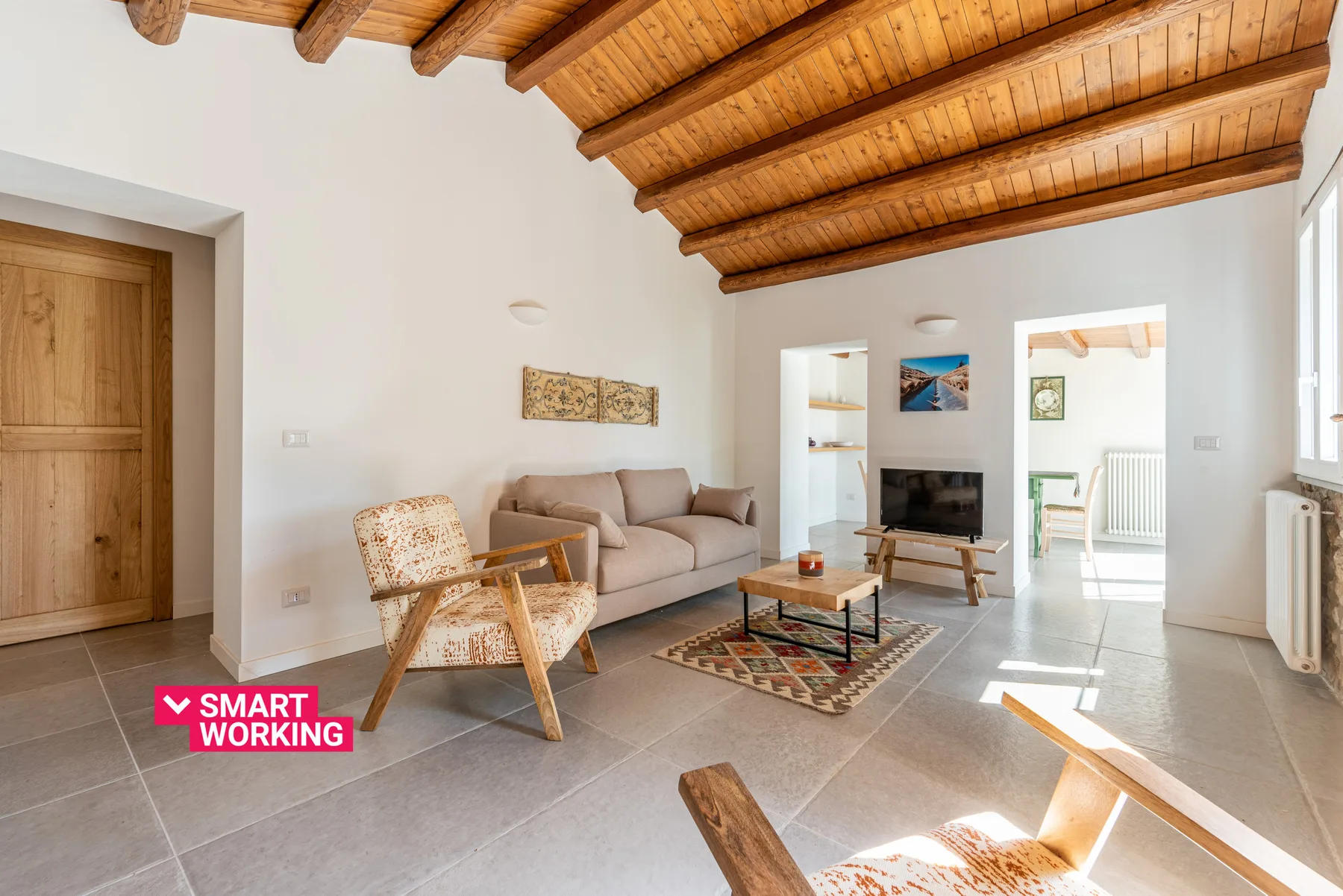 A bright living room with seating, a television, and exposed wooden ceiling beams.