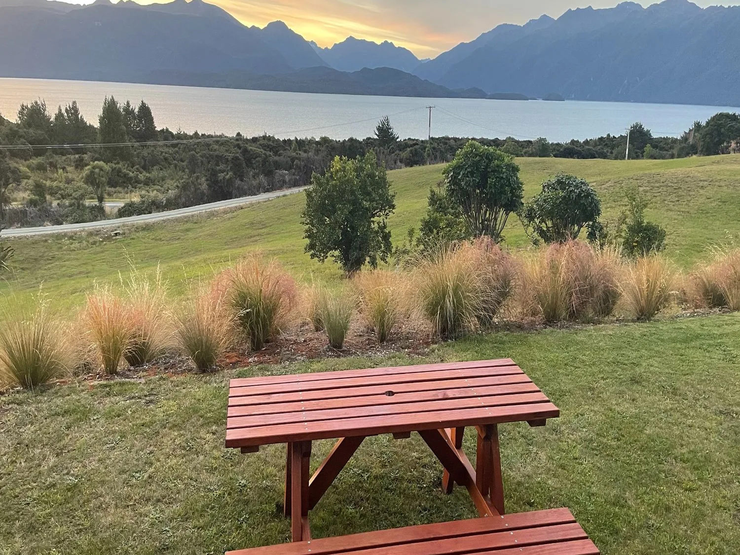 Una mesa de pícnic mira hacia un paisaje cubierto de hierba, un lago y montañas.