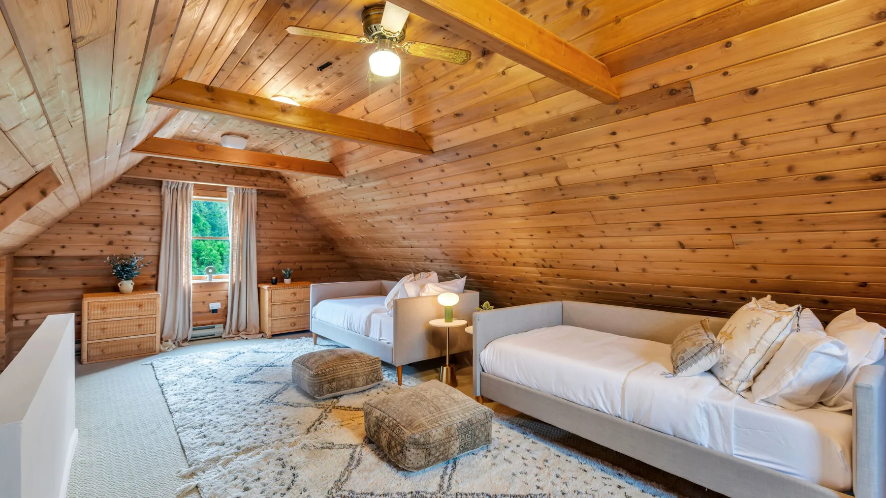 Un dormitorio abuhardillado con paredes de madera, dos camas, una alfombra estampada y un ventilador de techo.