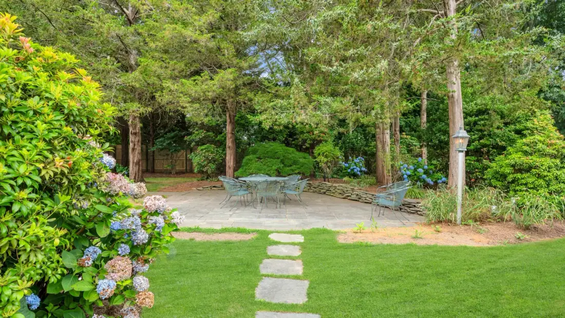 Un patio de jardín con asientos al aire libre está rodeado de árboles, arbustos floridos y césped.