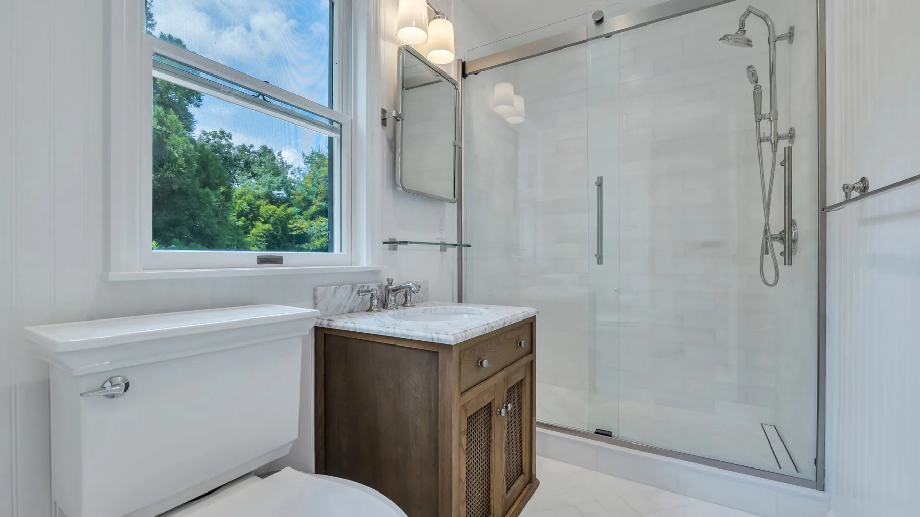 Un baño luminoso con ducha de vidrio, tocador de madera y ventana con vistas a la vegetación.