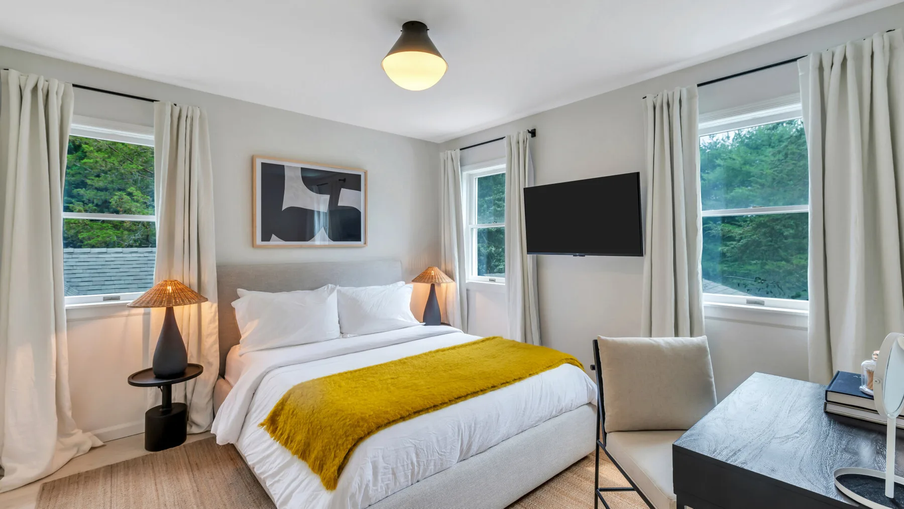 Un dormitorio con una cama, una manta color mostaza y ventanas con cortinas.